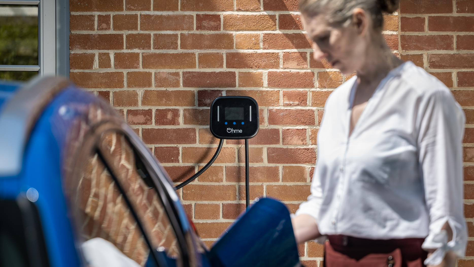 Person standing next to a blue EV with the Ohme Home Pro on the wall behind