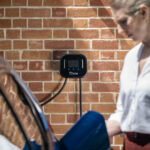 Person standing next to a blue EV with the Ohme Home Pro on the wall behind